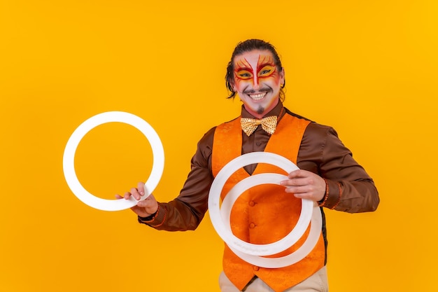 Portrait of a juggler in a vest with a painted face juggling hoops on a yellow background