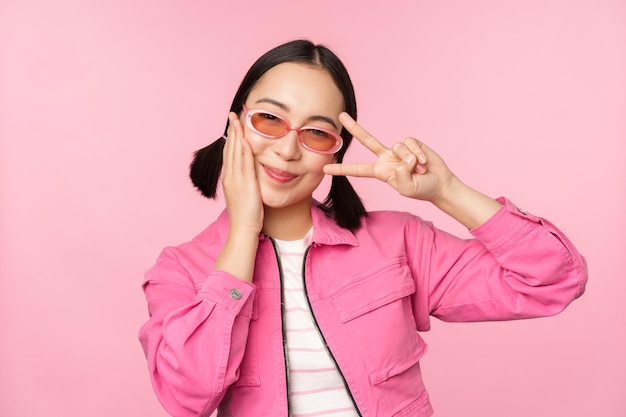 Portrait of kawaii asian girl in sunglasses showing peace vsign near eye and smiles cute at camera posing in trendy clothes against pink background