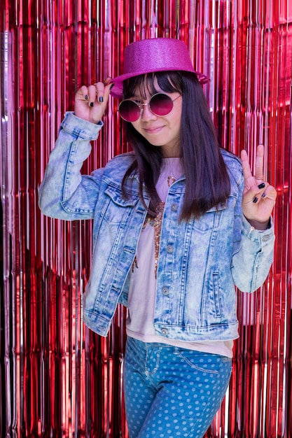 Portrait of a latin teen girl with a hat and sunglasses at a party making a sign of love and peace with her fingers