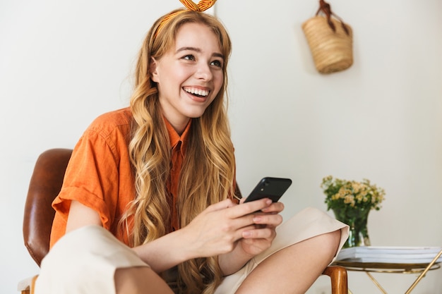 Portrait of a laughing cute happy young blonde woman at home indoors using mobile phone sitting on chair.