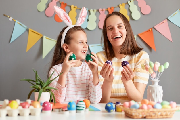 Photo portrait of laughing extremely happy mother and daughter wearing bunny ears holding colored eggs celebrating easter at home posing against gray decorated wall