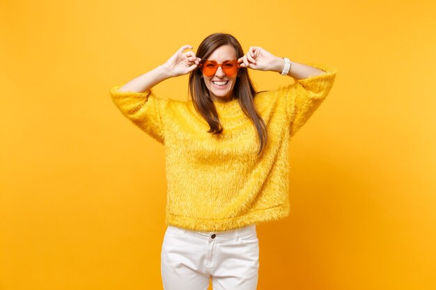 Photo portrait of laughing happy young woman in fur sweater, white pants holding heart orange eyeglasses isolated on bright yellow background. people sincere emotions, lifestyle concept. advertising area.