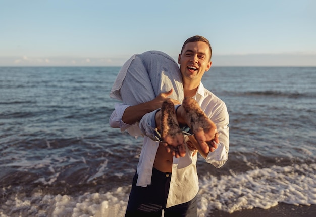 Portrait of laughing man holding woman on his shoulder standing on beach by sea