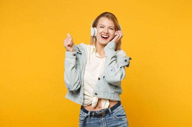 Portrait of laughing young woman in denim casual clothes listen music with headphones, looking camera isolated on yellow orange wall background in studio. People lifestyle concept. Mock up copy space.