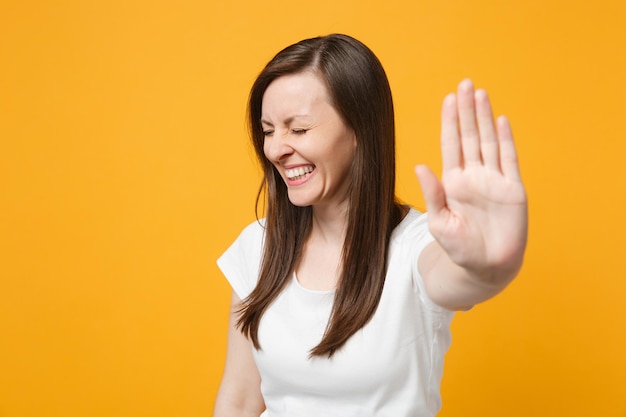 Portrait of laughing young woman in white casual clothes keeping eyes closed, showing palm on camera isolated on yellow orange wall background in studio. People lifestyle concept. Mock up copy space.