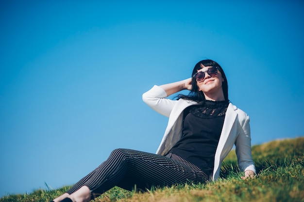 Portrait of a life style brunette girl in sunglasses on the grass in the park in a white jacket.