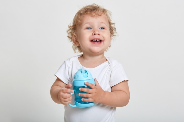 Portrait of little blond boy, dressed in white shirt, posing isolated over light wall with blue bottle for baby food, wants drinking water, looking away with excited expression.