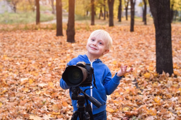 Portrait of a little blond boy with a large DSLR camera on a tripod Photo session in the autumn park