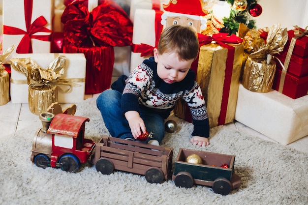 Portrait of little blonde boy sitting on the floor in decorated studio and playing with Christmas presents and boxes