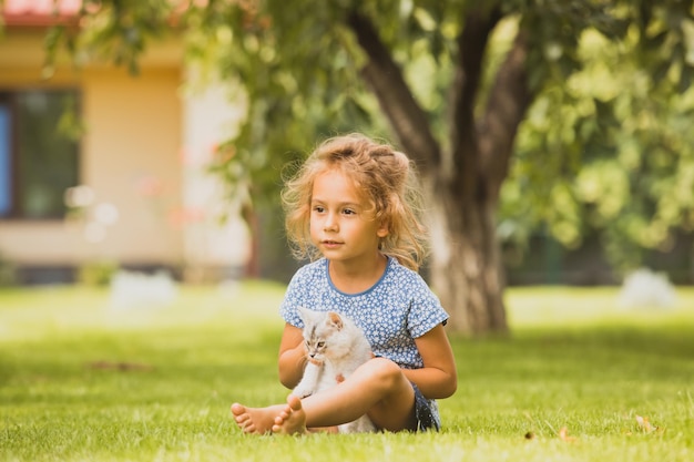 Portrait of a little blonde girl who is sitting with a kitten on a green lawn The cute girl hold a pet in the backyard