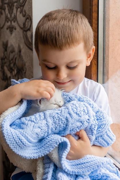 Portrait of a little boy holding a white kitten wrapped in a blue knitted blanket in his hands