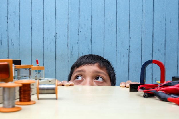 Portrait of little boy looking from under the table with STEM project on the table. Schoolboy who is