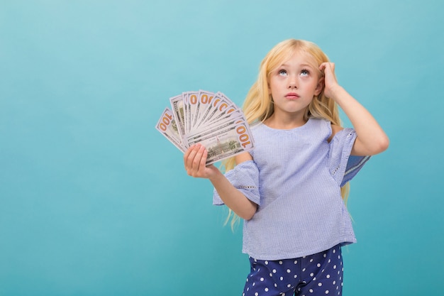 Portrait of little caucasian girl in blue t-shirt with long blonde hair holds money isolated on blue