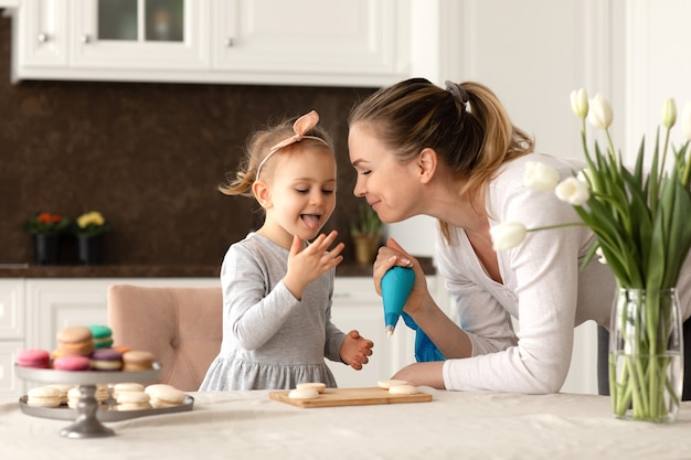 Portrait of little funny girl and her mother baking macarrons and cookies in the kitchen. Happy family and mothers day concept