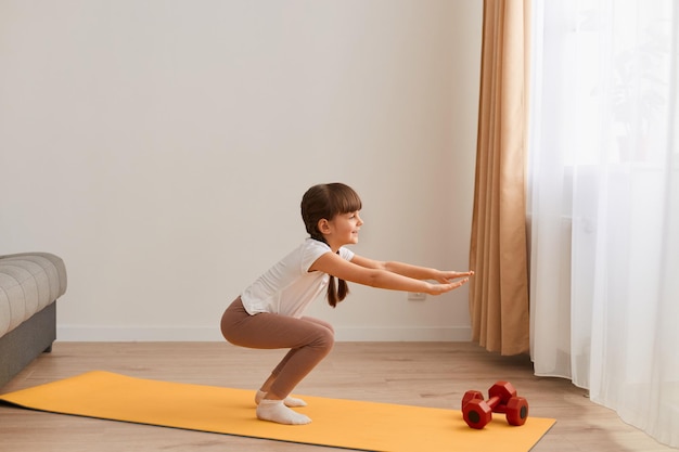 Portrait of little girl doing squat exercises workout at home, cute kid training on a mat indoor, dark-haired female kid having exercises near the window in her room.