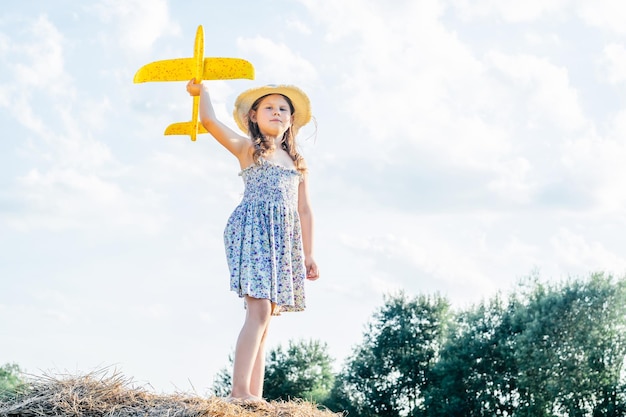 Portrait of little girl in hat and dress playing flying yellow toy airplane standing on haystack Taking aim in sky