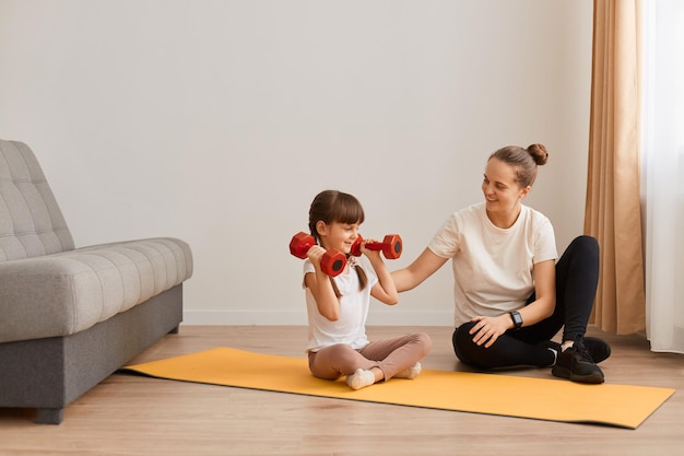 Portrait of little girl holding red dumbbells in hands, mother teaching her daughter to do sport exercises, sitting on floor on yoga mat, fitness at home, children healthy lifestyle.