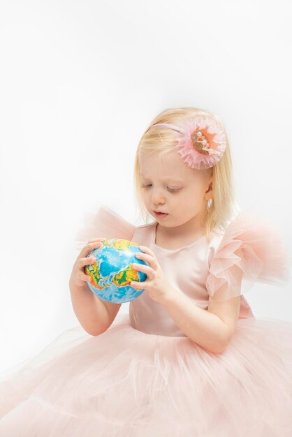 Portrait of little girl in pink dress with decoration hoop holds globe model in hands Blonde girl white background