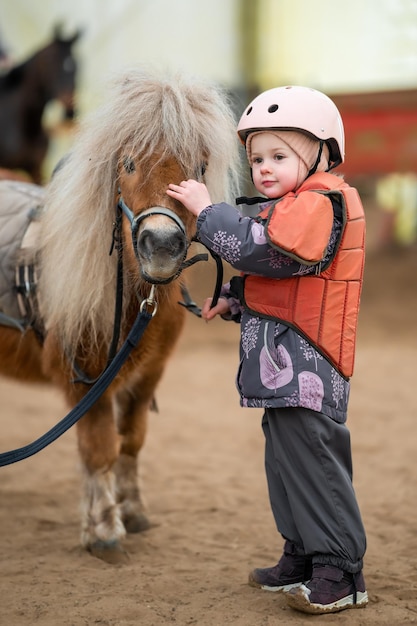 Portrait of little girl in protective jacket and helmet with her brown pony before riding lesson