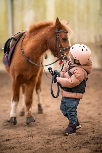 Portrait of little girl in protective jacket and helmet with her brown pony before riding lesson