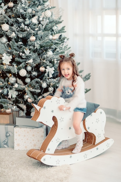 Portrait of a little girl sitting on a wooden horse against the background of a Christmas tree