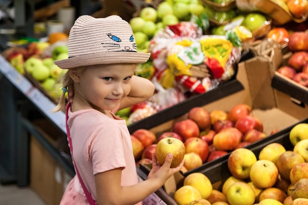 Portrait little girl with apple standing in grocery food fruit store deep thinking and looking at camera Adorable cute kid boy fresh fruits