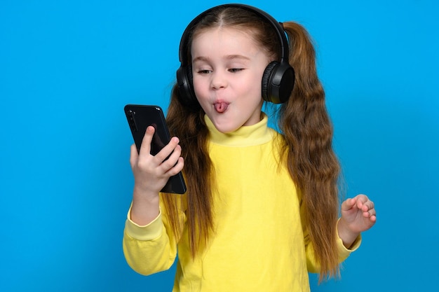 Portrait of a little girl with a phone and wireless headphones a child in a yellow sweater on a blue background