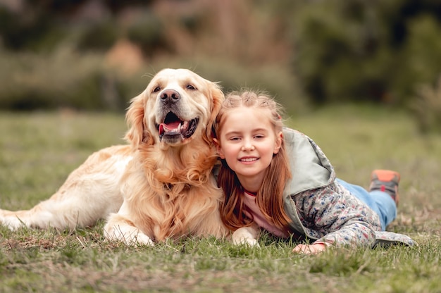 Portrait of little happy girl lying on the grass with golden retriever dog in spring time