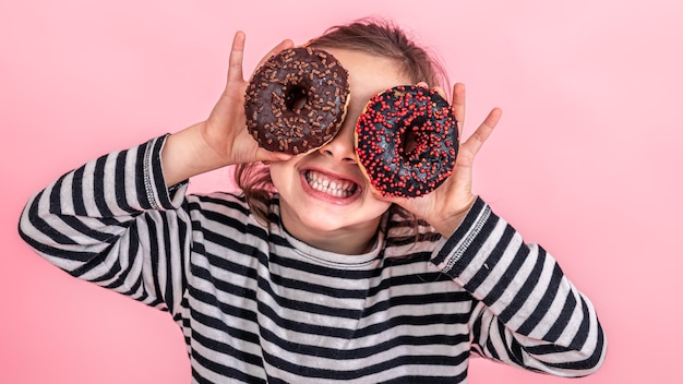 Portrait of a little smiling girl brunette with and two appetizing donuts in her hands, closes her eyes with donuts, on a pink background.