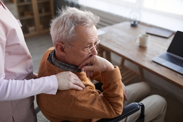 Portrait of lonely senior man in wheelchair at retirement home with nurse comforting him, copy space