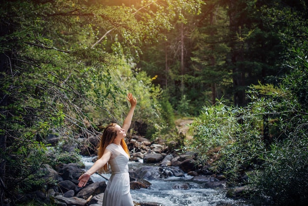 Portrait long-haired brunette in white dress posing against small mountain river and green trees. Beautiful young woman on the forest stream shore.