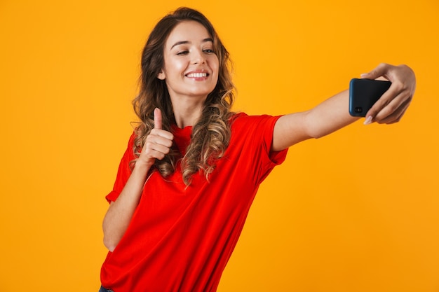 Portrait of a lovely cheerful young woman standing isolated over yellow wall, taking a selfie