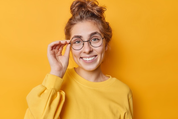 Portrait of lovely European woman keeps hand on rim of spectacles smiles gladfully looks self confident at camera wears casual jumper has pleasant conversation isolated over yellow background