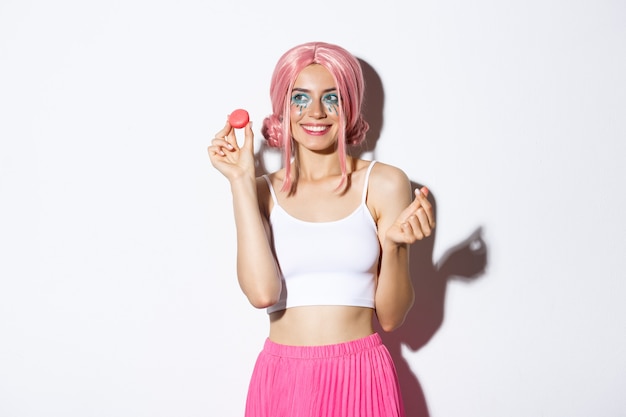 Portrait of lovely female model eating macaroons and smiling, wearing pink wig and outfit for party, standing over white background