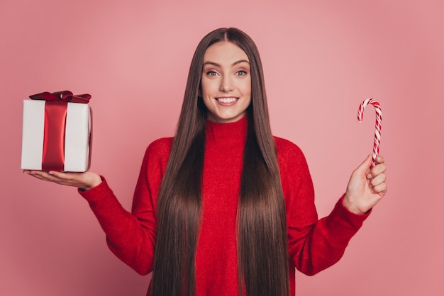 Portrait of lovely girl holding xmas candy stick big giftbox isolated over pink background