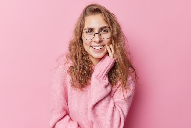 Portrait of lovely positive woman with long wavy hair smile toothily keeps hand on cheeks feels glad to have pleasant conversation wears long sleeved jumper round spectacles poses against pink wall