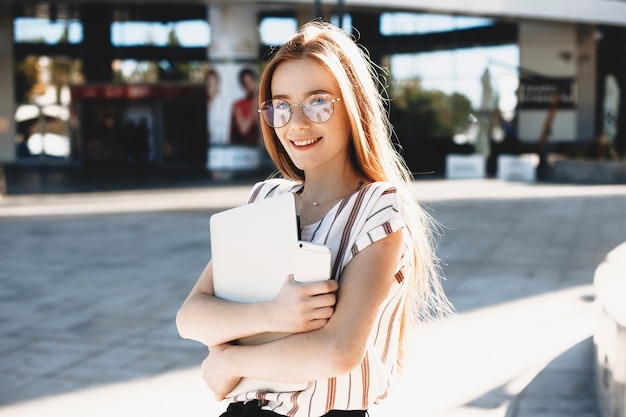 Portrait of a lovely red haired female student with freckles looking at camera smiling and holding a smartphone and a laptop against a building.