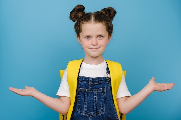 Portrait of lovely sweet small schoolgirl wears yellow backpack raise two palms arms holding empty space beaming shiny smiling positive good look, posing isolated over blue color background in studio