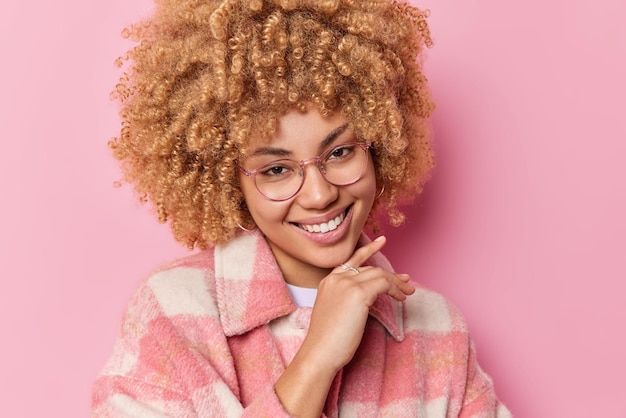 Portrait of lovely woman has natural curly hair touches jawline gently smiles toothily looks happily at camera dressed in fashionable coat isolated over pink background Pleasant feelings concept