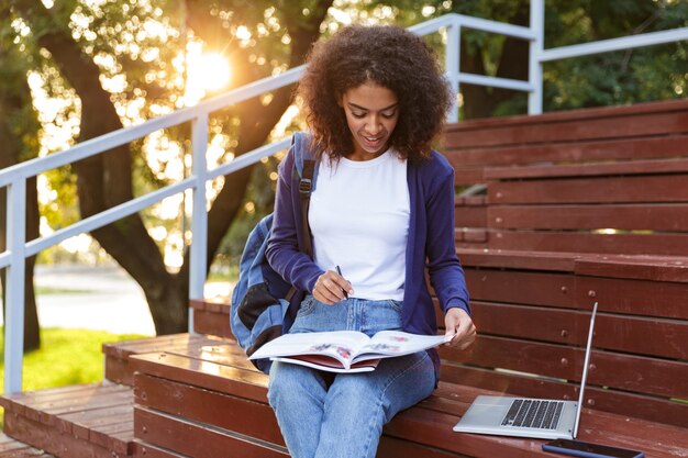 Portrait of a lovely young african girl with backpack resting at the park, reading magazine