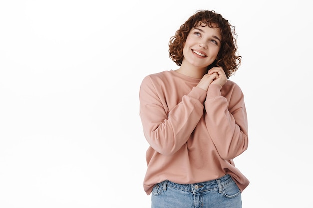 Portrait of lovely young girl daydreaming, looking at top with love and romantic emotion, holding hands together and gazing up, mesmerized by something beautiful, white background.