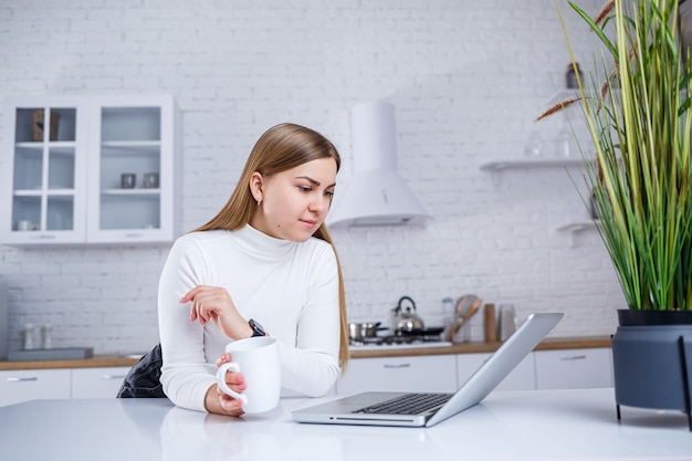 Portrait of lovely young woman studying at home on laptop. A girl with long blond hair in a sweater drinks tea. Modern kitchen interior and education concept. Work remotely from home