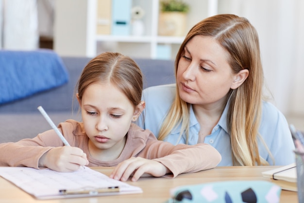 Portrait of loving adult mother looking at cute little girl doing homework or test while studying at home in cozy interior