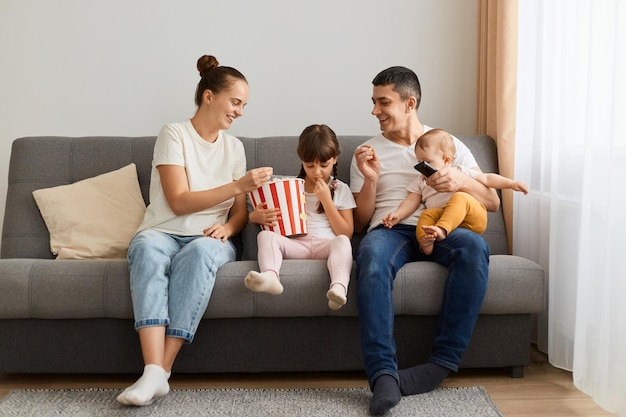 Portrait of loving couple sitting on sofa with their kids and having fun while watching funny video or films enjoying merry weekend at home eating popcorn