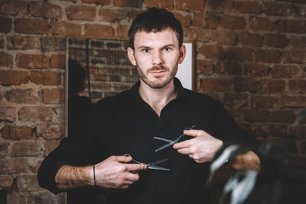 Photo portrait male barber holds sharp scissors against a brick wall background. barbershop advertising concept. place for text or advertising