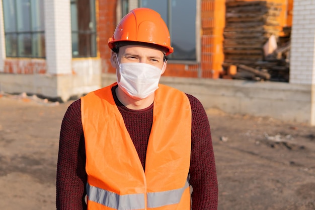 Portrait of male construction worker in medical mask and overalls on background of house under construction