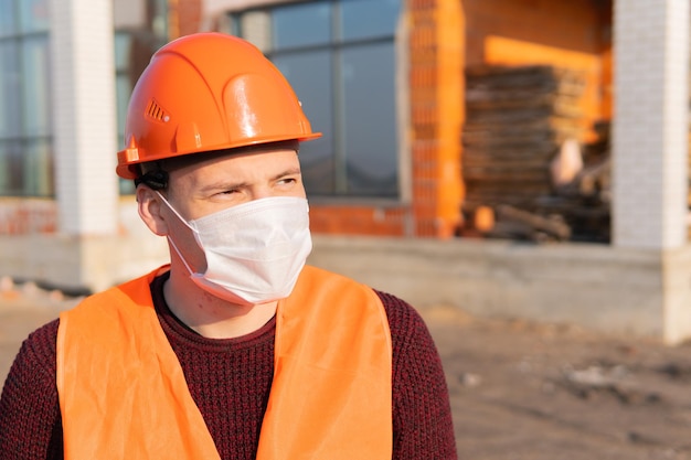 Portrait of male construction worker in medical mask and overalls on background of house under construction