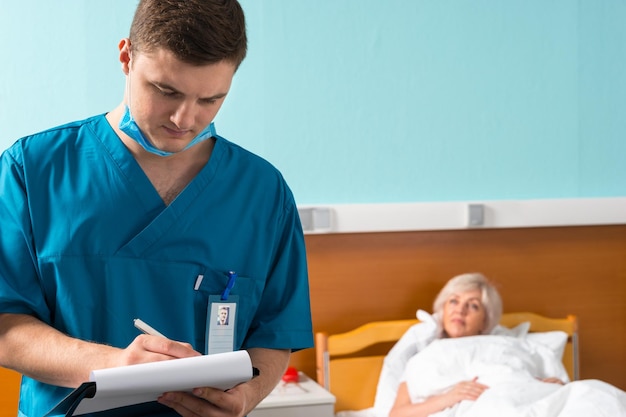 Portrait of male doctor in uniform wearing a mask making some notes in report at clipboard, ill patient lying in the hospital bed in the background. Healthcare concept