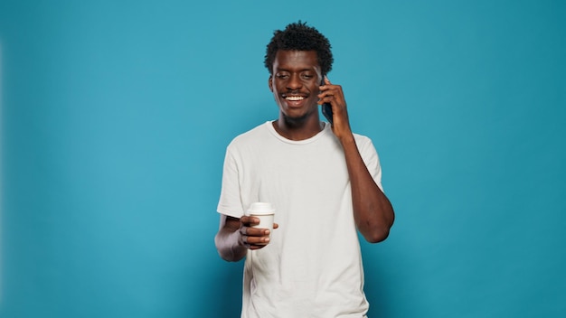 Portrait of man answering phone call on smartphone in studio while holding cup of coffee. Person using mobile phone for remote communication, smiling and talking on touchscreen device