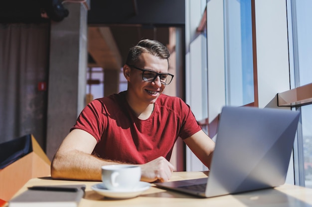 Portrait of a man IT professional working remotely with a modern laptop sitting at a table and smiling at the camera during a break a happy human programmer in vision correction glasses